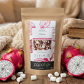 Sachet d'infusion Pitaya Papothe en papier kraft posé sur une table en bois, entouré de fruits du dragon frais roses et de mini-chamallows blancs. Ambiance cocooning chaleureuse avec arrière-plan flou de plaids en maille et lumière douce.